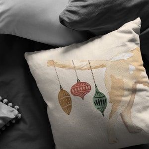May include: A white pillow with a watercolor illustration of a branch with three ornaments hanging from it. The ornaments are red, green, and gold.