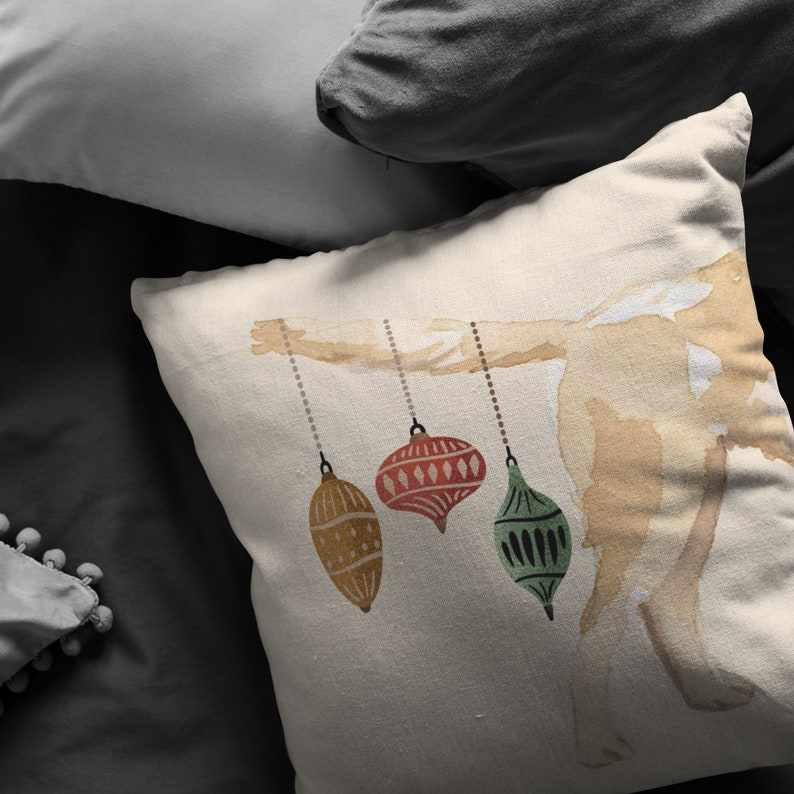 May include: A white pillow with a watercolor illustration of a branch with three ornaments hanging from it. The ornaments are red, green, and gold.