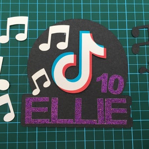 May include: A black and white TikTok logo with a red and blue outline, surrounded by white musical notes. The name "ELLIE" is written in purple glitter letters with the number "10" in purple glitter next to it.