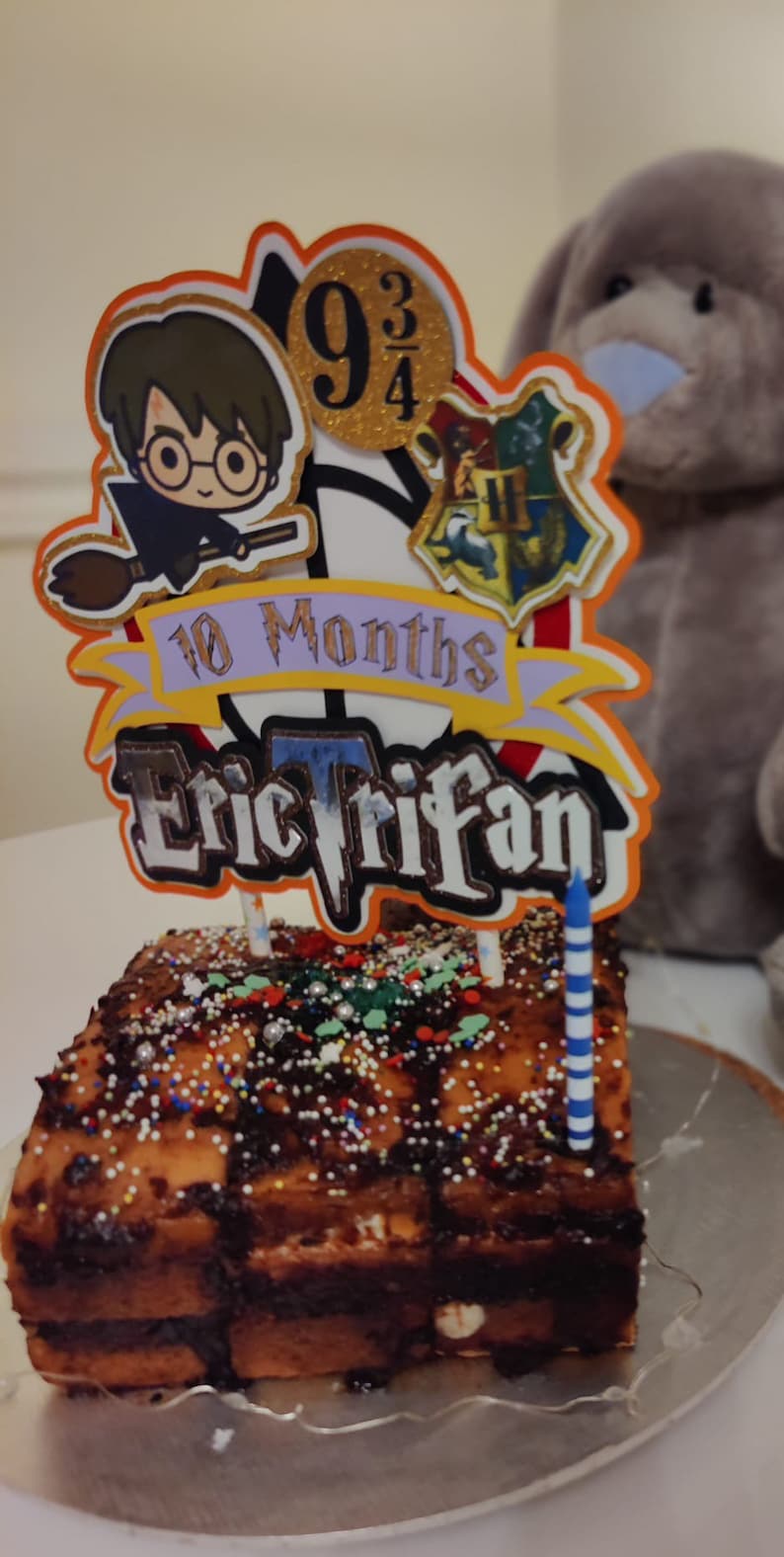 May include: A chocolate cake with sprinkles and a Harry Potter themed cake topper. The topper features a character from the Harry Potter series, the number 9 3/4, and the Hogwarts crest. The cake topper also includes the text "10 Months Eric Trifan".