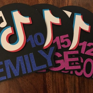 May include: Three black and white TikTok logo party decorations with glitter text. The text reads "Emily 10", "Age 15", and "12 years old".