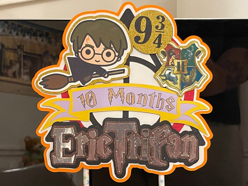 May include: A Harry Potter themed cake topper with a cartoon character holding a broom, a Hogwarts house crest, and the text "9 3/4" and "10 Months Eric Trifan".