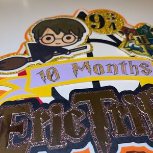May include: A Harry Potter themed cake topper with a cartoon image of Harry Potter flying on a broomstick. The topper also includes a banner that says "10 Months" and the name "Eric" in gold glitter letters. The topper is decorated with gold glitter and a Hogwarts house crest.