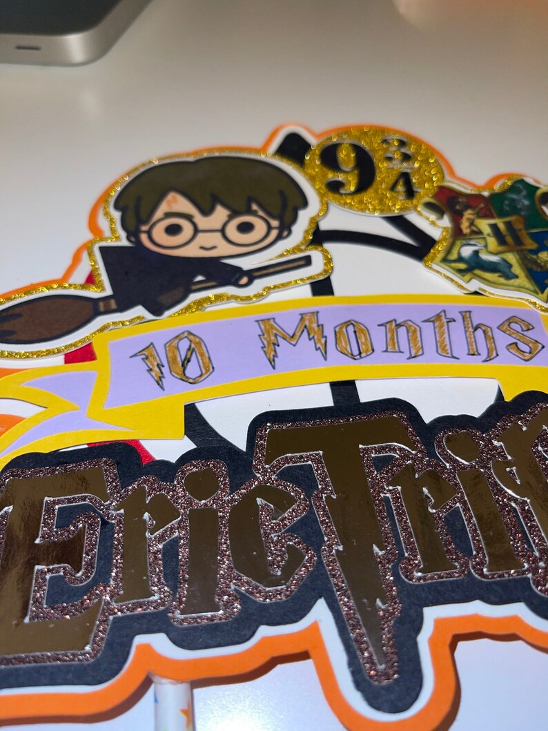May include: A Harry Potter themed cake topper with a cartoon image of Harry Potter flying on a broomstick. The topper also includes a banner that says "10 Months" and the name "Eric" in gold glitter letters. The topper is decorated with gold glitter and a Hogwarts house crest.