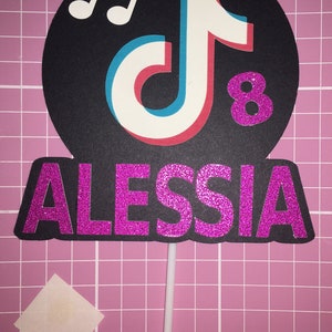 May include: A black and white TikTok logo with a musical note above it. The number 8 is in pink glitter next to the logo. The name ALESSIA is in pink glitter below the logo. The logo is on a white stick.