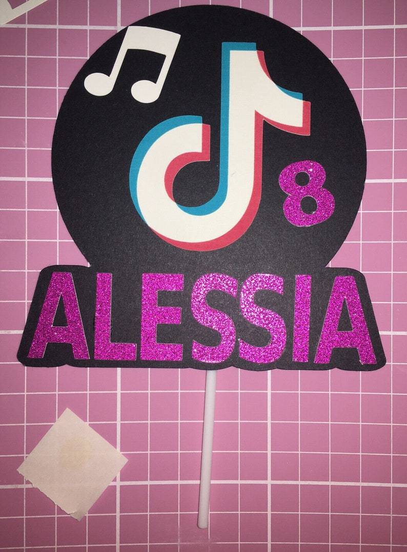 May include: A black and white TikTok logo with a musical note above it. The number 8 is in pink glitter next to the logo. The name ALESSIA is in pink glitter below the logo. The logo is on a white stick.