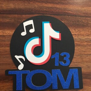 May include: Black and blue glitter cake topper with a white, blue, and red TikTok logo, two musical notes, and the number 13. The name "TOM" is written below the logo.