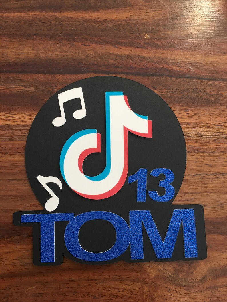 May include: Black and blue glitter cake topper with a white, blue, and red TikTok logo, two musical notes, and the number 13. The name "TOM" is written below the logo.