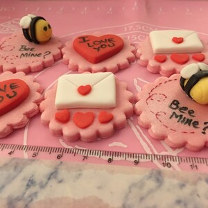 May include: Six pink and white fondant cupcake toppers with Valentine's Day themes. The toppers feature a heart with the text "I love you", a white envelope with a red heart, and a bee with the text "Bee Mine?"