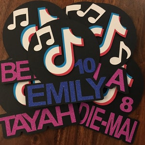 May include: A set of six black and white TikTok-themed party decorations. The decorations feature the TikTok logo, musical notes, and the names "TAYAH", "EMILY", "DIE-MAI", "BE", and "A". The numbers "10" and "8" are also included.