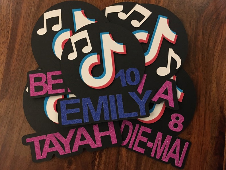 May include: A set of six black and white TikTok-themed party decorations. The decorations feature the TikTok logo, musical notes, and the names "TAYAH", "EMILY", "DIE-MAI", "BE", and "A". The numbers "10" and "8" are also included.