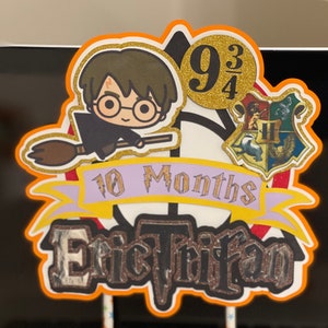 May include: A Harry Potter themed cake topper with a cartoon image of Harry Potter flying on a broomstick. The topper also includes a Hogwarts house crest, a banner that says "10 Months", and a gold circle with the number "9 3/4". The name "Eric Trifan" is written in black with glitter on a black background.