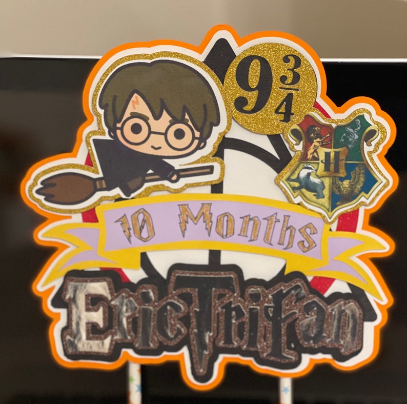May include: A Harry Potter themed cake topper with a cartoon image of Harry Potter flying on a broomstick. The topper also includes a Hogwarts house crest, a banner that says "10 Months", and a gold circle with the number "9 3/4". The name "Eric Trifan" is written in black with glitter on a black background.