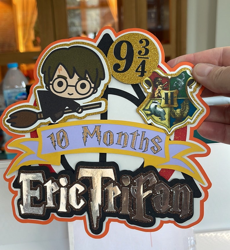 May include: A Harry Potter themed cake topper with a cartoon image of Harry Potter on a broomstick, a Hogwarts house crest, and the text "9 3/4", "10 Months", and "Eric Trifan".