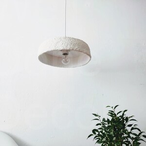 May include: A white, round, textured pendant light fixture with a single light bulb hanging from a white ceiling.