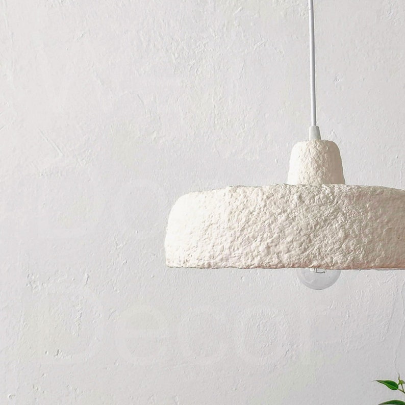 May include: A white paper mache pendant light fixture hangs from a white cord against a white wall.