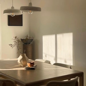 May include: A light wood dining table with six chairs, a white vase with dried branches, and two small terracotta bowls on a white napkin. The table is illuminated by two white pendant lights with exposed bulbs.