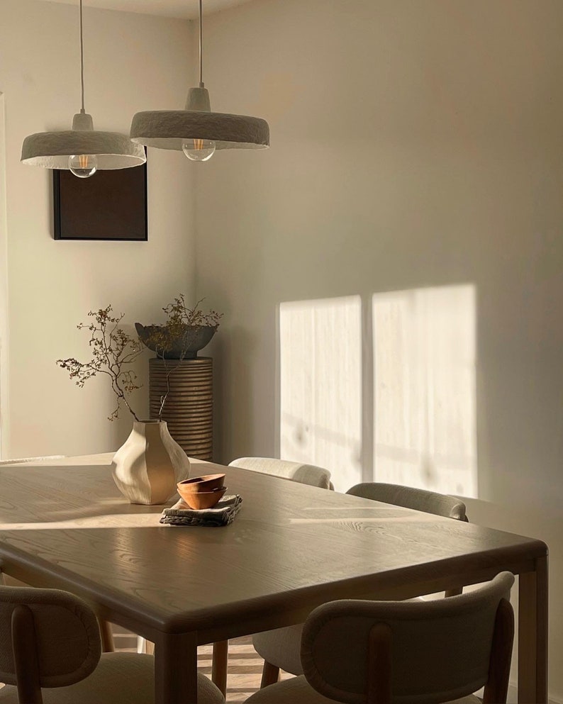 May include: A light wood dining table with six chairs, a white vase with dried branches, and two small terracotta bowls on a white napkin. The table is illuminated by two white pendant lights with exposed bulbs.