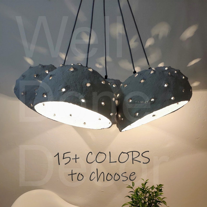May include: A set of five gray paper mache pendant lights with holes throughout the surface. The lights are hanging from black cords and are illuminated. The text "15+ COLORS to choose" is written below the lights.