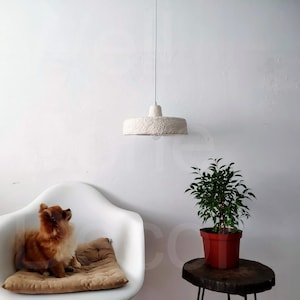 May include: A white, fluffy, paper lampshade hangs from the ceiling above a white armchair with a brown cushion. A small potted plant with green leaves sits on a wooden table.