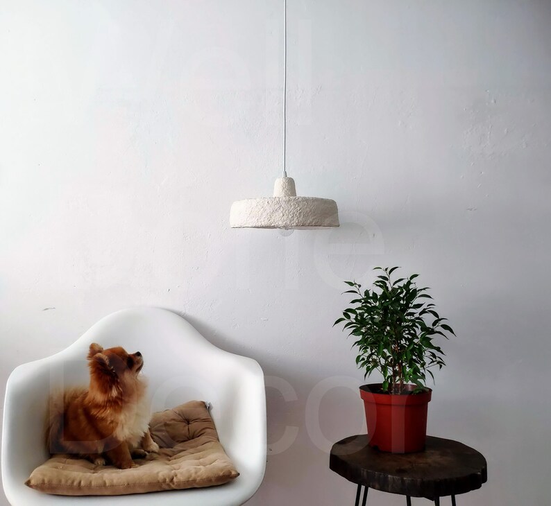 May include: A white, fluffy, paper lampshade hangs from the ceiling above a white armchair with a brown cushion. A small potted plant with green leaves sits on a wooden table.