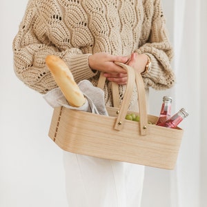 May include: A woman is holding a wooden picnic basket with a baguette, grapes, and bottles of pink beverage inside. The basket has two handles and is made of light brown wood.