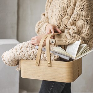 May include: A light brown knitted blanket is placed in a wooden basket with a natural finish. The basket has two handles and is filled with books.