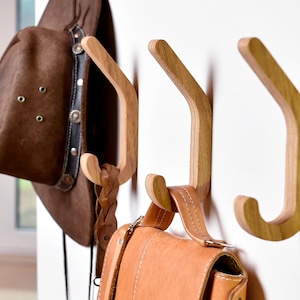 Könnte beinhalten: Drei Wandhaken aus Holz mit geschwungenem Design. Ein brauner Hut und eine braune Ledertasche hängen an den Haken.