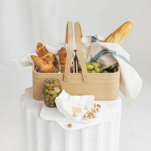 May include: A wooden picnic basket with a blue and white striped cloth, filled with croissants, olives, grapes, and a block of brie cheese. The basket is sitting on a white speckled surface.