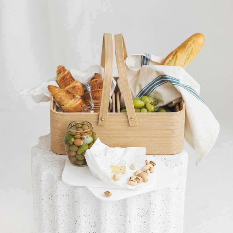 May include: A wooden picnic basket with a blue and white striped cloth, filled with croissants, olives, grapes, and a block of brie cheese. The basket is sitting on a white speckled surface.