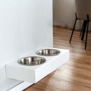 May include: White wall-mounted pet food and water bowl stand with two stainless steel bowls. The stand is mounted to a white wall and is in a modern kitchen.