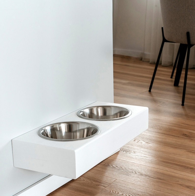 May include: White wall-mounted pet food and water bowl stand with two stainless steel bowls. The stand is mounted to a white wall and is in a modern kitchen.