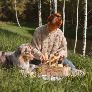 May include: A woman in a beige sweater sits on a blanket in a forest setting with a dog and a picnic basket filled with bread. She is holding a knife and cutting the bread.