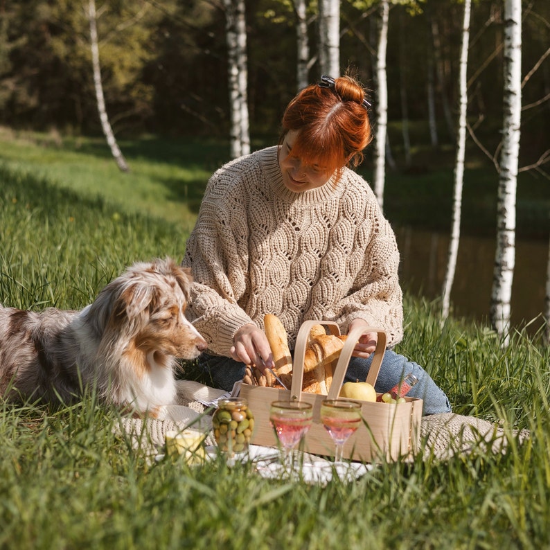 May include: A woman in a beige sweater sits on a blanket in a forest setting with a dog and a picnic basket filled with bread. She is holding a knife and cutting the bread.