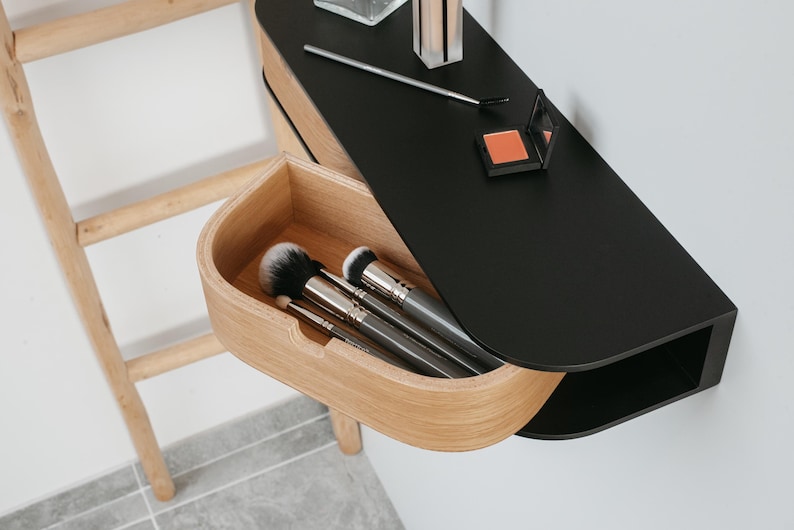 May include: A black wall-mounted shelf with a wooden drawer that is open, revealing makeup brushes. The shelf has a curved edge and a small compartment on the side.