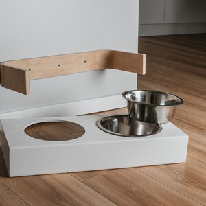 May include: White elevated dog bowl stand with two stainless steel bowls. The stand is mounted to a wall with a wooden shelf.