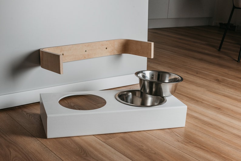 May include: White elevated dog bowl stand with two stainless steel bowls. The stand is mounted to a wall with a wooden shelf.
