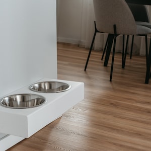 May include: White wall-mounted pet food and water bowl stand with two stainless steel bowls. The stand is mounted on a white wall and is in a room with hardwood floors.