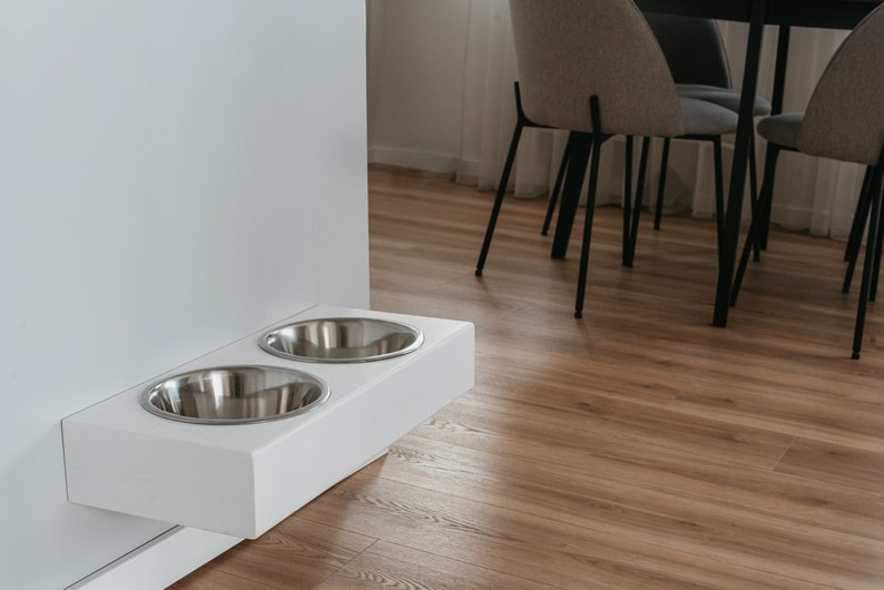 May include: White wall-mounted pet food and water bowl stand with two stainless steel bowls. The stand is mounted on a white wall and is in a room with hardwood floors.