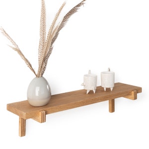 May include: A wooden shelf with a light brown finish and two white ceramic candle holders. A vase with dried brown feathers is on the shelf.