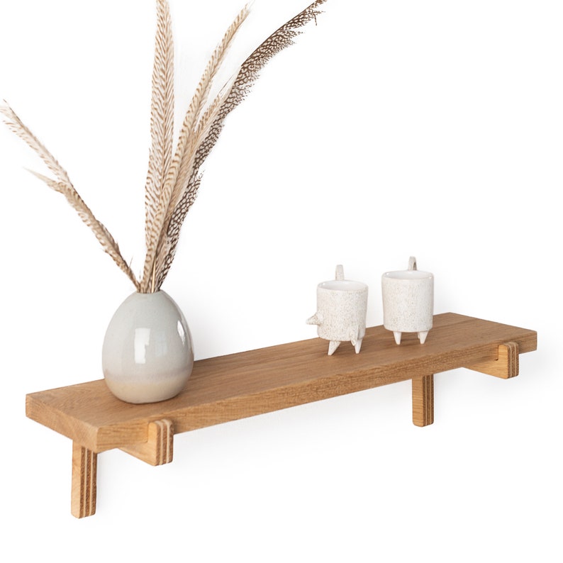 May include: A wooden shelf with a light brown finish and two white ceramic candle holders. A vase with dried brown feathers is on the shelf.