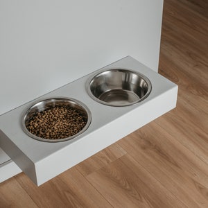 May include: White wall-mounted pet food and water bowl set with two stainless steel bowls. One bowl is filled with brown pet food.