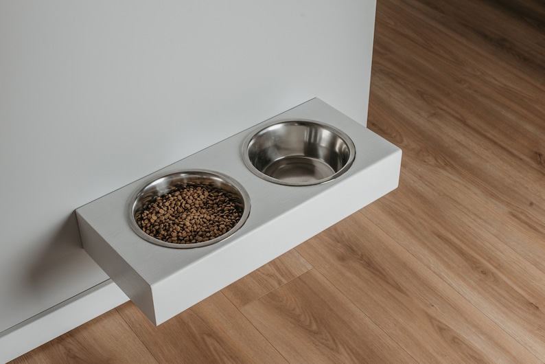 May include: White wall-mounted pet food and water bowl set with two stainless steel bowls. One bowl is filled with brown pet food.