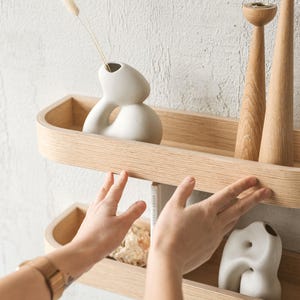 May include: Two wooden shelves with rounded edges are mounted on a white wall. The shelves are filled with decorative items, including two white ceramic vases, two wooden candle holders, and a book. The shelves are made of light wood and have a natural finish.