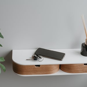 May include: A white wall-mounted shelf with two drawers, a black smartphone, keys, and a black glass diffuser with wooden sticks.