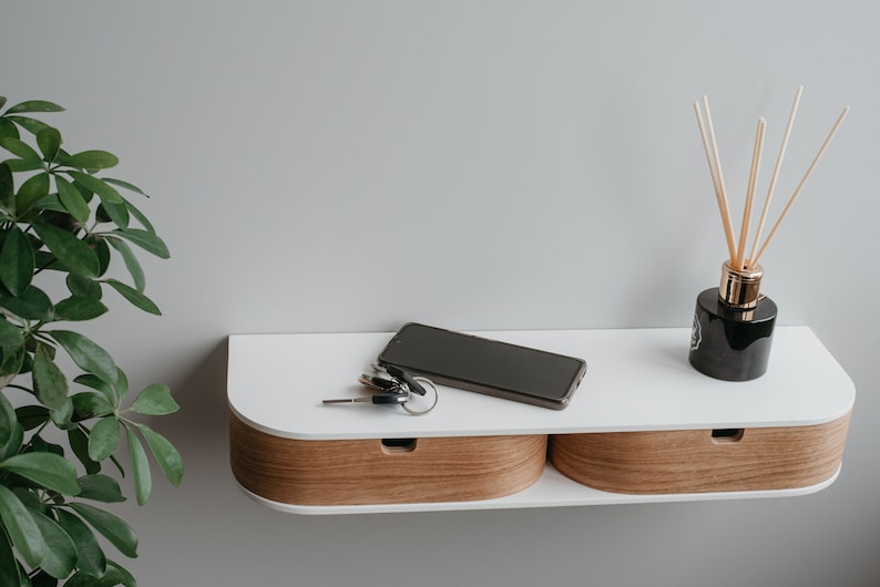 May include: A white wall-mounted shelf with two drawers, a black smartphone, keys, and a black glass diffuser with wooden sticks.