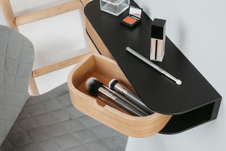 May include: A black wall-mounted vanity with a wooden drawer. The drawer is open and contains makeup brushes. The vanity also has a shelf with a bottle of foundation, a compact, and a makeup brush.