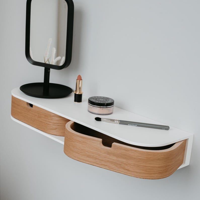 May include: White wall-mounted shelf with two drawers. The shelf has a curved front edge and is made of wood. The drawers are made of wood with a natural finish. There is a small round jar of face powder, a lipstick, a makeup brush, and a small black mirror on the shelf.