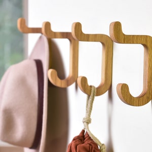 May include: Four wooden wall hooks in a J shape, mounted on a white wall. A hat and a small bag are hanging on the hooks.