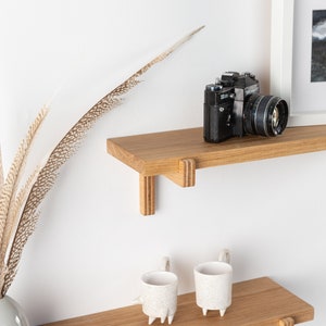 May include: Two wooden shelves with a vintage camera on the top shelf and two white ceramic mugs on the bottom shelf. The shelves are mounted on a white wall.